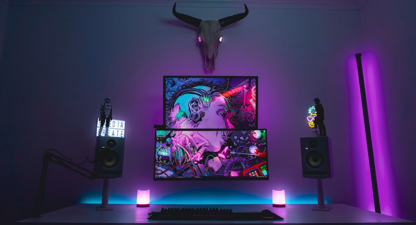 A neon-lit gaming setup with two stacked monitors showing colourful artwork, flanked by speakers and accent lighting.