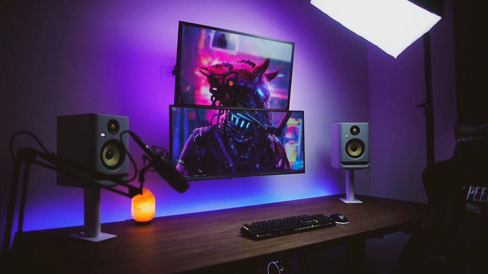 A purple-lit gaming desk with two stacked monitors, studio speakers, a microphone arm and a backlit keyboard.